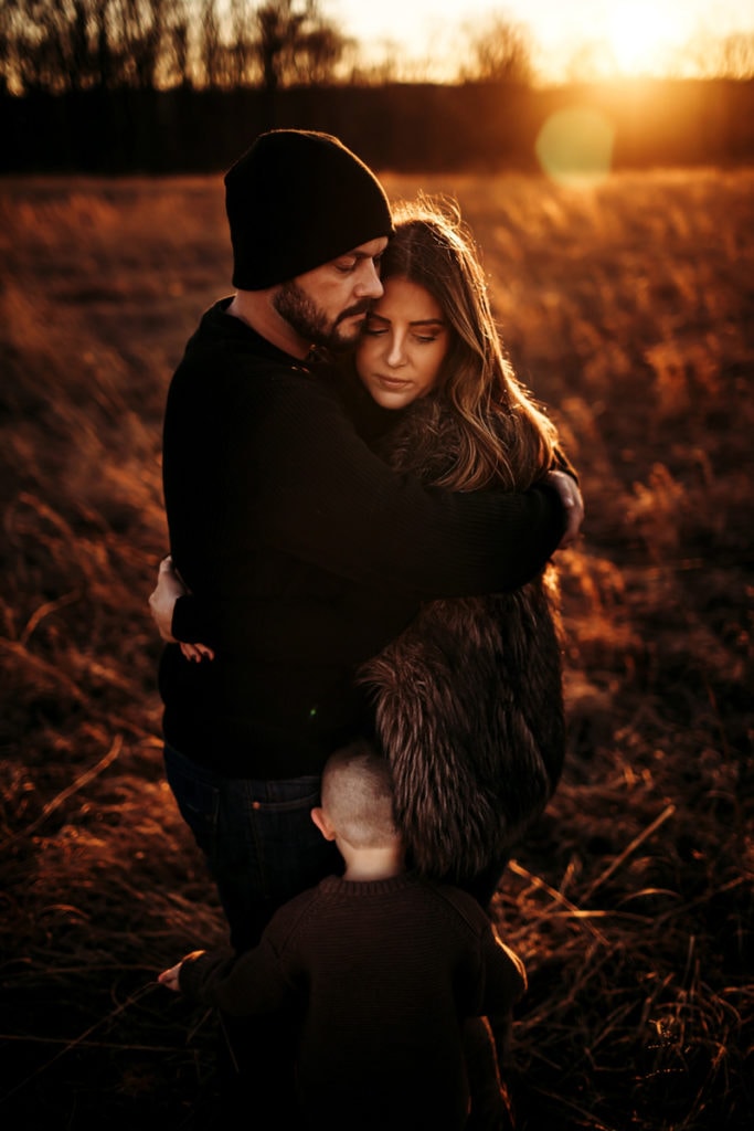 Family Photographer, a husband and wife embrace outside at golden hour, their young son hugs them too