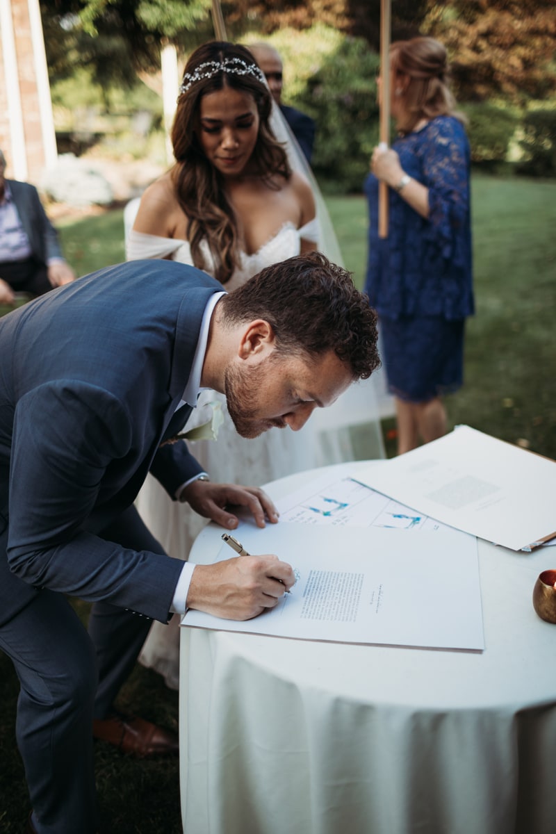 Wedding Photographer, groom signs wedding certificate at wedding ceremony as bride looks on