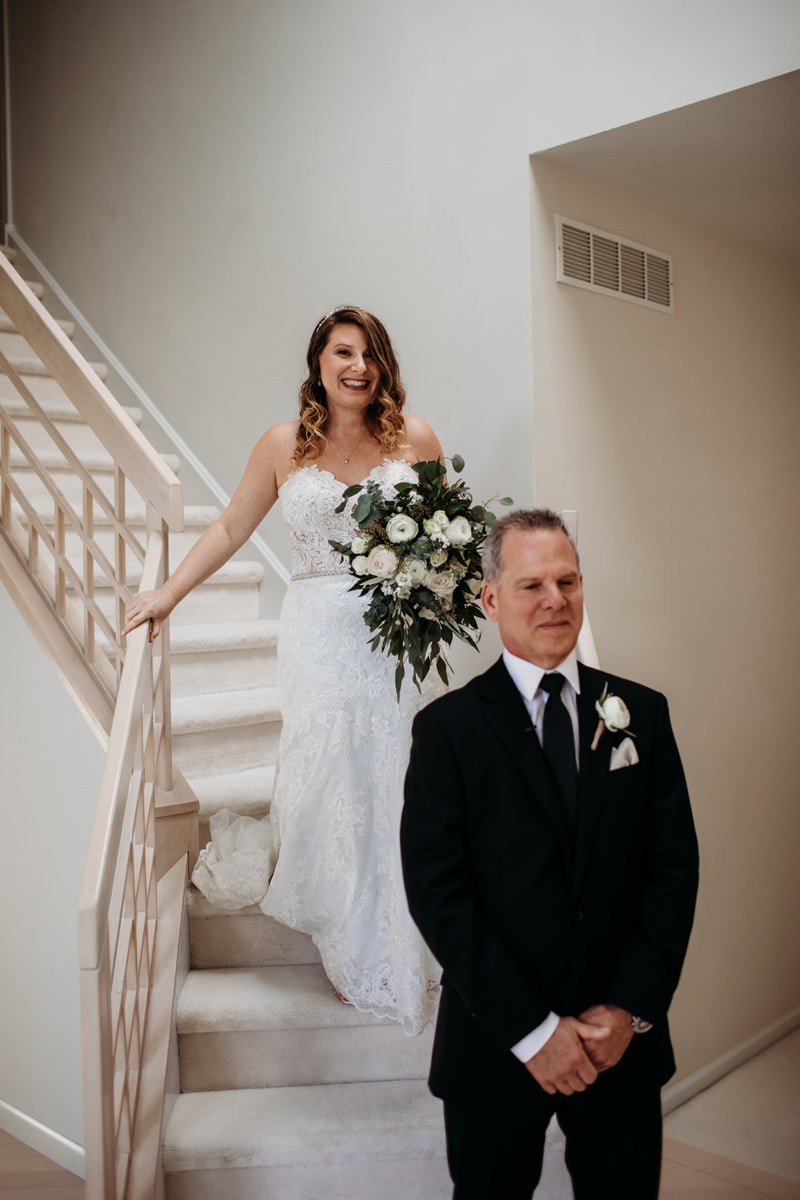 Wedding Photographer, a father stands before the stairs as his daughter, the bride, is about to surprise him in her dress